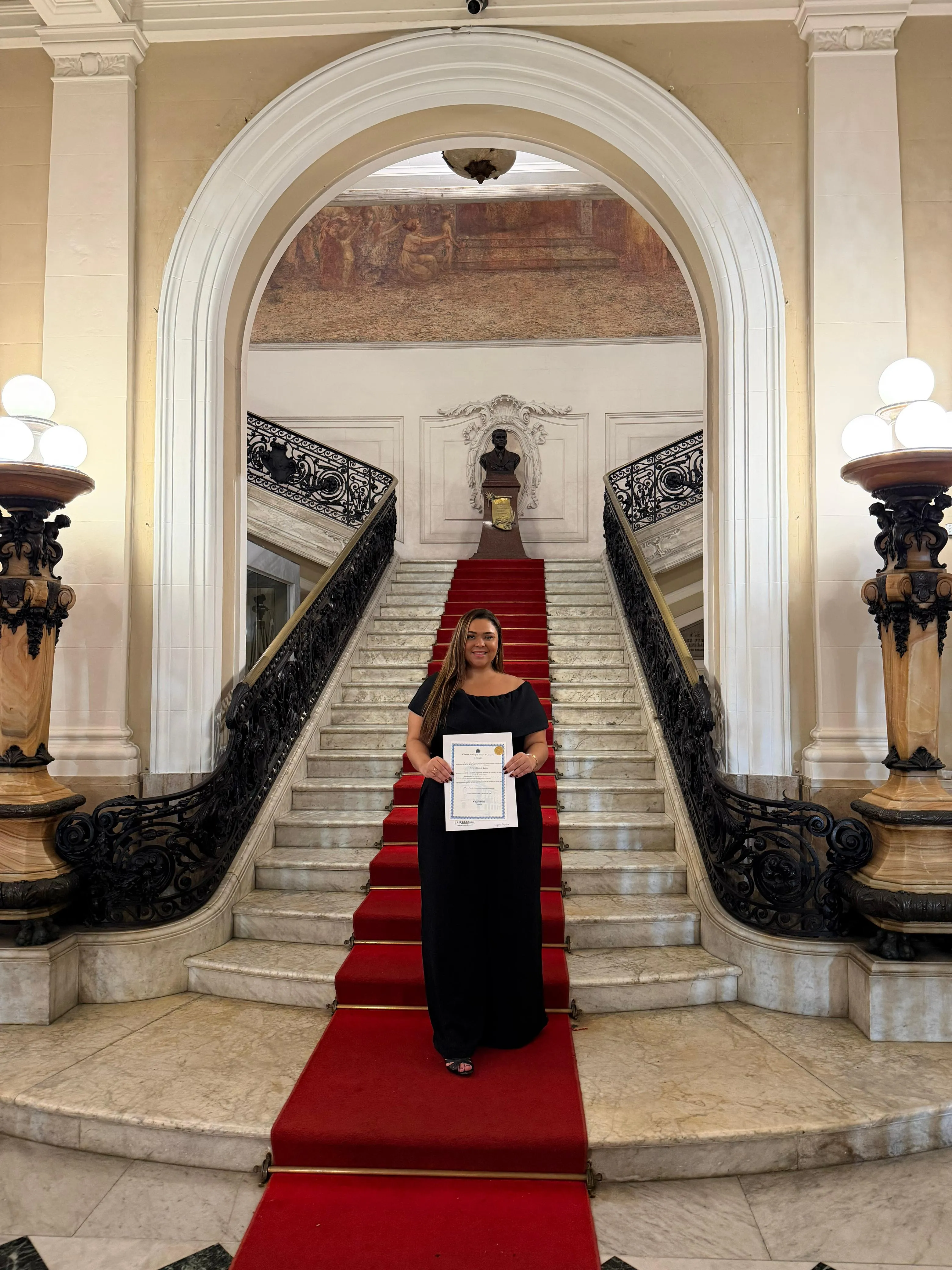 Dra. Nayara Ribeiro na escadaria histórica da Câmara Municipal do Rio de Janeiro segurando sua Moção de Reconhecimento